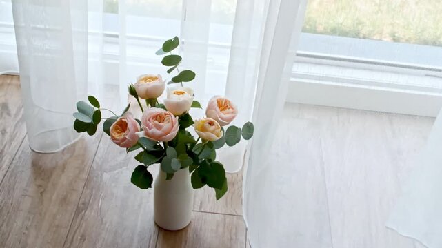Roses and eucalyptus flowers stand in a vase by the window. Wind blows through the open window in the room. Waving white tulle near the window. Product presentation, mockup for advertising. 4k