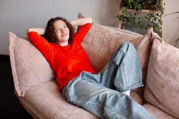 Obraz premium Woman relaxing on sofa in living room. Teenage girl sitting comfortably on couch at home with hands behind head. Leisure, peace and serenity. Lifestyle concept of rest, comfort.