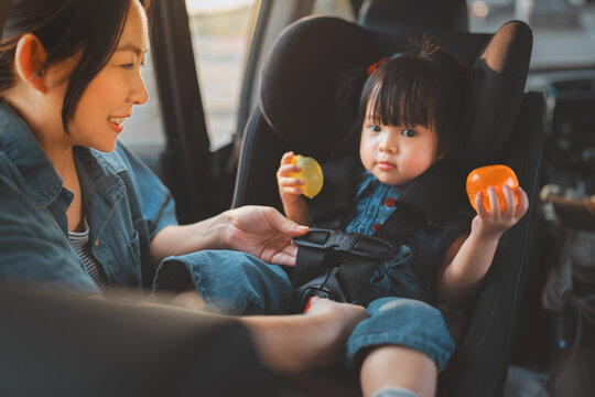 A loving Asian mother carefully buckles her smiling toddler daughter into a secure car seat before a road trip. Concept of child safety, parenting, and family travel security