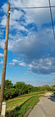 Obraz premium A tall wooden utility pole stands beside a narrow asphalt road winding through the green hills of Gornje Psarjevo village under a cloudy blue sky
