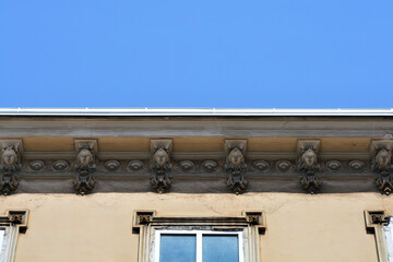 Decorative sculpted stone faces adorn the beige facade under the roof gutter of an old classic building against a blue sky © nahhan