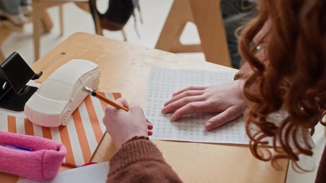 High angle over the shoulder shot of unrecognizable girl with curly red hair checking answers on paper during test at desk in modern classroom
