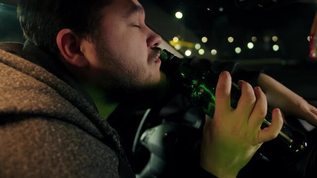 Portrait of isolated drunk caucasian bearded man drinks alcohol in car at night. The tired driver sits in the car and drinks from a green bottle of whiskey. Antisocial behavior and illegal activities