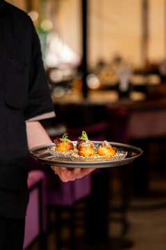 A waiter in a black shirt holds a silver tray with gourmet appetizers served on small glass plates in a restaurant.