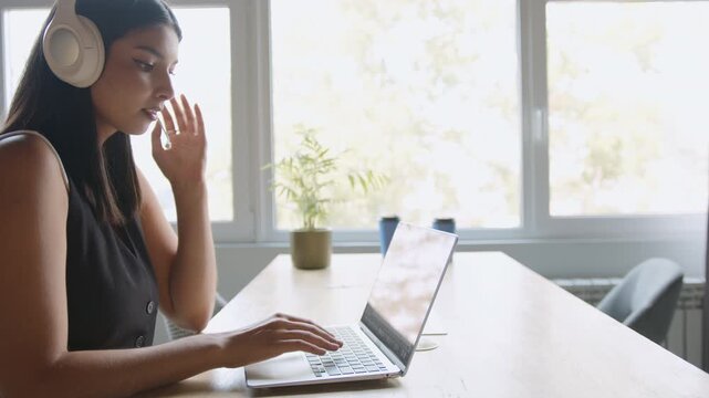 An attentive Indian customer service agent effectively communicates with clients via a headset, actively typing on a laptop to address inquiries from a brightly lit contemporary office
