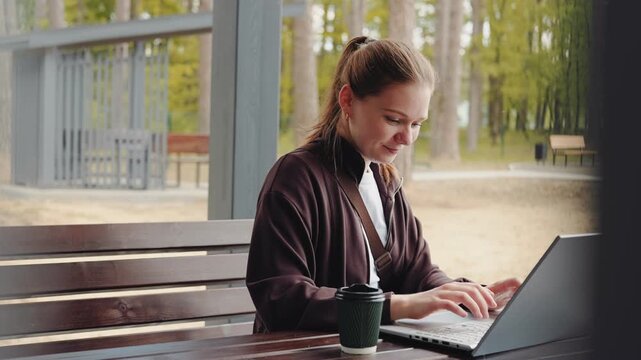 A young woman types on her laptop while seated at a wooden table in an outdoor area with trees around her. She enjoys a drink while focusing on her work