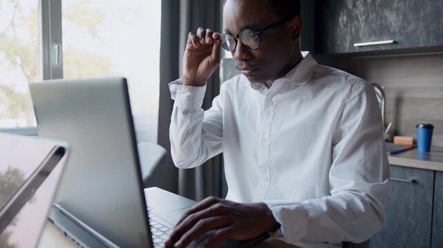 A Black man in a white shirt and glasses is intensely working on his laptop during the day in a modern home office. He adjusts his eyewear, showing focus and dedication to his professional tasks