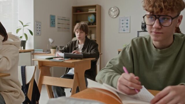 Student wearing glasses and green sweatshirt intently writing in notebook against other students at their desks in modern classroom