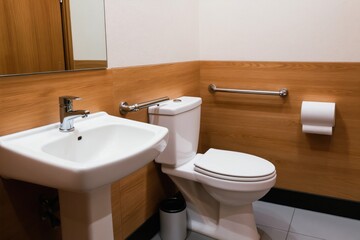 Fototapeta premium Clean modern restroom with white porcelain toilet and sink. Grab bars offer support for elderly or disabled persons. Wood accent walls add warmth to the sanitary space.