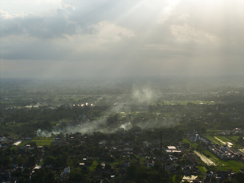 Aerial view of lush green fields and residential areas with smoke rising under a dramatic sky with sunbeams in Kecamatan Prambanan, Daerah Istimewa Yogya, Indonesia.