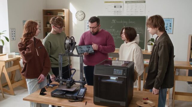 Teacher holding digital tablet to show students 3D models of gears at table equipped with two 3D printers in modern classroom
