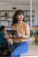 Fototapeta premium Diverse coworkers with Hispanic woman sitting on desk edge holding tablet in open-plan office