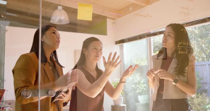 Diverse female team gathering at glass board by tablet, gesturing, writing with marker, finalizing