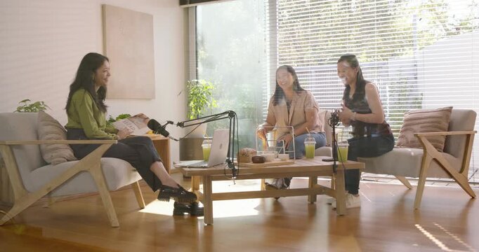 Three women hosting podcast, Asian host referencing notebook sparking guests gesturing at table mic