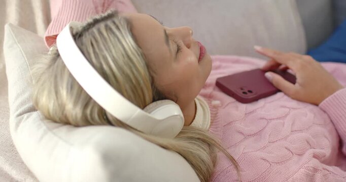 African American woman reclining on sofa, noticing maroon phone, tapping, smiling with headphones