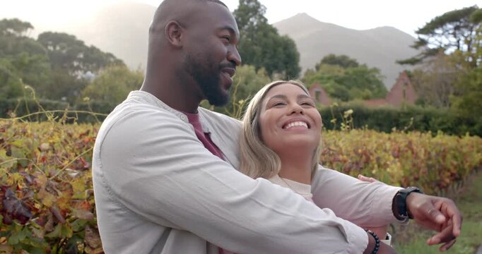 Diverse couple standing in vineyard embracing and sharing view, man pointing after spotting house
