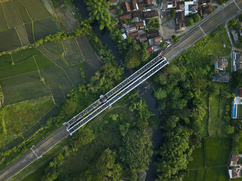 Aerial view of a railway bridge crossing a river surrounded by green paddy fields and lush vegetation in Kecamatan Prambanan, Daerah Istimewa Yogya, Indonesia.