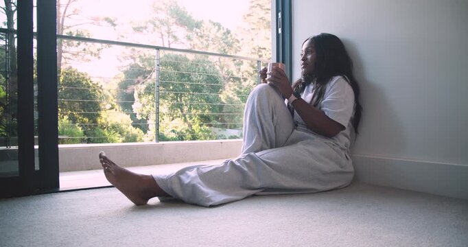 Adult African woman sitting by open sliding door, holding mug, gazing at sunlit-trees, copy space