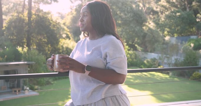 African woman leaning on balcony railing holding mug, reacting to camera tightening, smiling