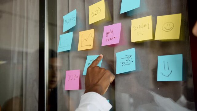 A hand points at bright sticky notes on a transparent board in an office, showcasing project tasks and data. Notes include numbers, percentages, and days of the week for planning and organization