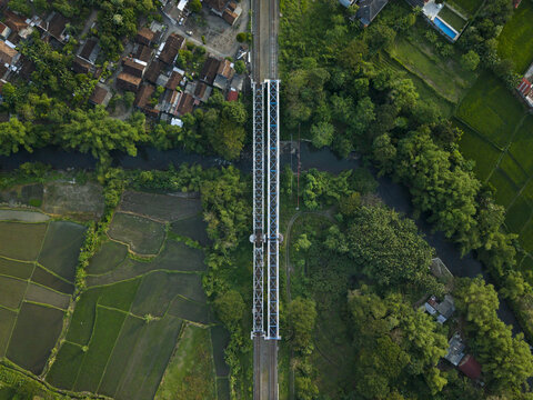 Aerial view of a railway bridge crossing a river surrounded by green rice paddies and lush trees in Kecamatan Prambanan, Daerah Istimewa Yogya, Indonesia.