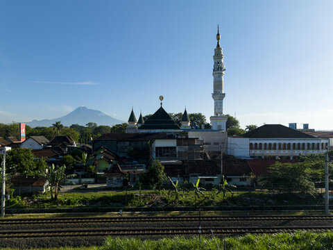 Aerial view of a mosque with a tall minaret and traditional rooftops with the Mount Merapi volcano in the background in Kecamatan Prambanan, Daerah Istimewa Yogya, Indonesia.