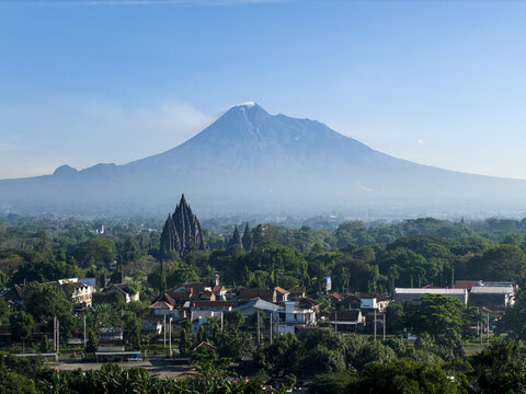 Aerial view of Prambanan Temple with Mount Merapi in the background, surrounded by lush greenery and residential rooftops in Kecamatan Prambanan, Daerah Istimewa Yogya, Indonesia.