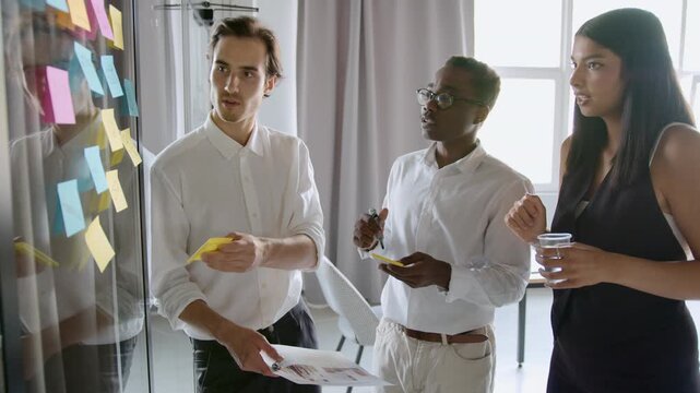 In an office, a diverse group of colleagues discusses ideas while standing by a wall filled with sticky notes. They share thoughts and take notes, working together to create new concepts