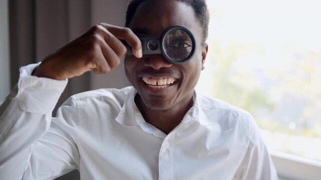 Cheerful black man smiles in an office, wearing a white shirt. He playfully holds a magnifying glass over one eye, humorously enlarging it while looking directly at the camera.