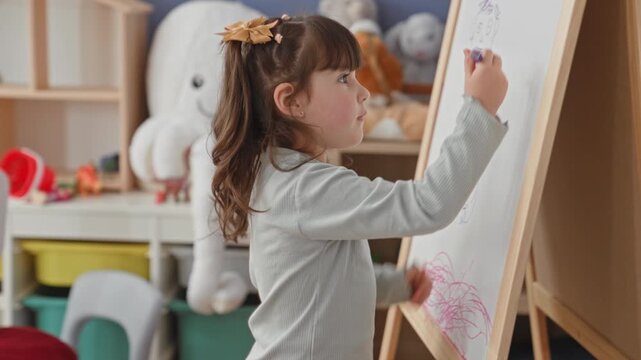 Girl drawing on wooden easel whiteboard with blue marker, right hand sketching a smiling face, side profile near large stuffed octopus in a home playroom building; creative curiosity.