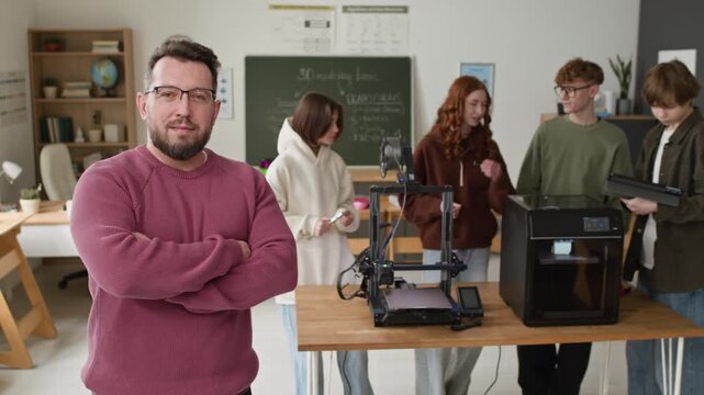 Portrait shot of male teacher wearing glasses and burgundy sweater posing with confident smile in bright 3D modeling classroom, where four students enthusiastically working with 3D printers