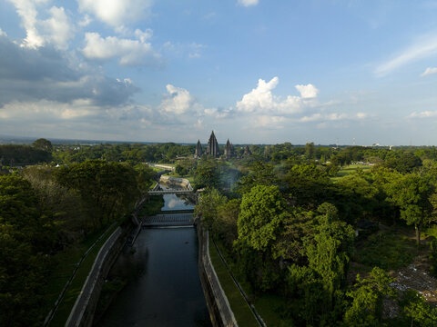 Aerial view of Prambanan Temple surrounded by lush tropical forests and a calm river under a blue sky with white clouds in Kecamatan Prambanan, Daerah Istimewa Yogya, Indonesia.