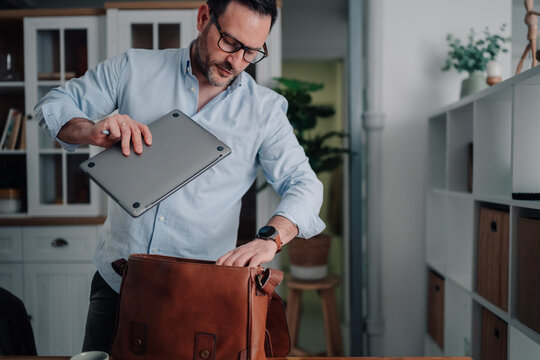 Man packing laptop into shoulder bag, preparing to leave home office for a remote work or travel destination