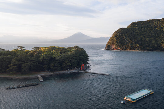 Aerial view of Mihama Beach and the red torii gate on the forested cape with Mount Fuji in the background under a soft sky Numazu, Shizuoka, Japan.