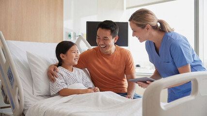 Fototapeta premium An Asian girl lies in a hospital bed smiling at her father and a Caucasian female nurse holding a digital tablet. A scene of professional medical care and family support in a modern clinic.
