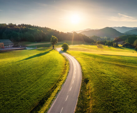 Aerial view of road in green meadows at sunset in summer. Top view from drone of rural road, mountains, forest. Beautiful landscape with roadway, trees, hills, fields, green grass, blue sky. Slovenia