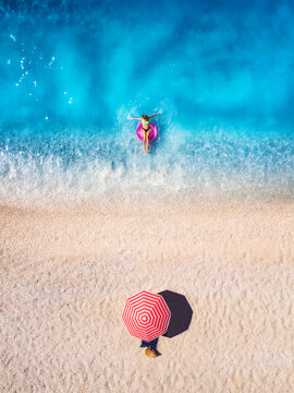 Aerial drone view of a woman swimming with pink swim ring in blue sea, sandy beach and red umbrella. Summer vacation and holiday. Tropical. Top down view of girl, clear water, waves. Lefkada, Greece
