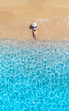 Aerial view of a lying woman in white hat on sandy beach and blue sea on sunny day. Summer vacation and holiday. Tropical. Top down view of girl, clear water, waves, sea coast. Sardinia island, Italy