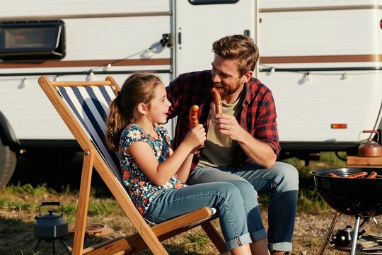 Father and daughter enjoy sausages from barbecue grill near camper van. Young family relax on deck chairs, eating food, having fun together outdoors on sunny day. Casual vacation lifestyle.