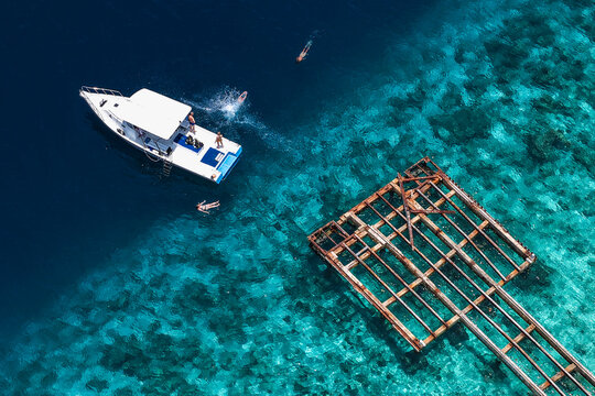 Aerial view of a white boat and people swimming near a rusty metal structure in clear turquoise water over coral reefs Muraidhoo, Haa Alif Atoll, Maldives.