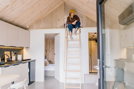 Young Woman Sitting In Cozy Scandinavian Tiny House Loft During Quiet Weekend Retreat