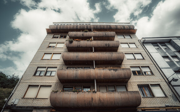 Low-angle view of a residential apartment facade in Spain, showing rounded weathered balconies, brick walls, shuttered windows and dramatic clouds above