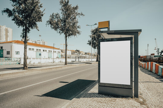 Blank ad panel on a modern bus shelter beside an empty road, ideal for outdoor campaign mock-up, with trees, sidewalks, warehouses and cranes in the distance; with a copy-space area on the left