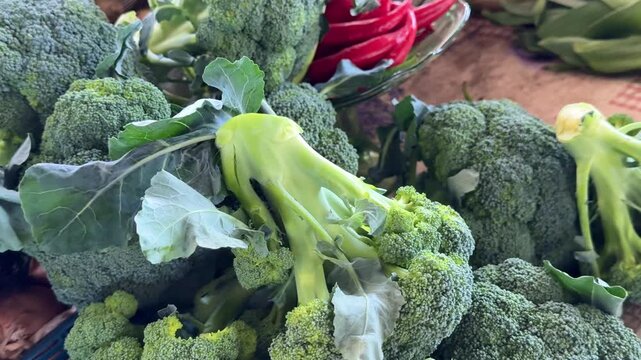 Green broccoli displayed on bazaar counter with fresh natural texture while bright red chili peppers softly visible in background creating colorful farm vegetable atmosphere.