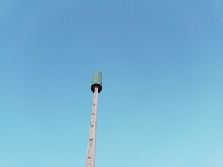 Fototapeta premium A tall amusement park drop tower ride against a clear blue sky, featuring a cylindrical passenger carriage at the top of a vertical steel structure with a ladder