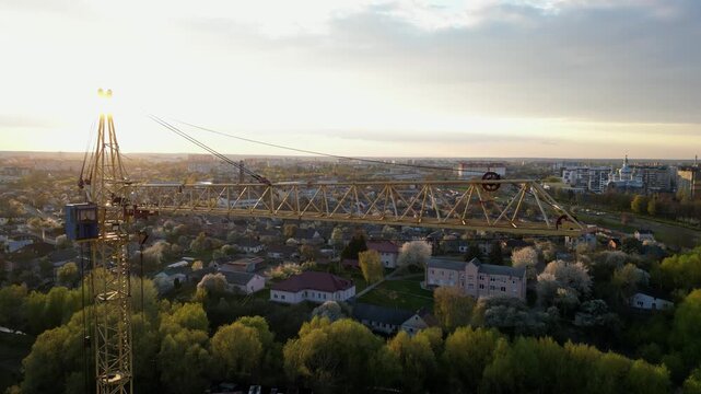Aerial footage showing a yellow tower crane on a construction site with a beautiful cityscape in the background during a golden sunset, symbolizing urban development and industrial progress