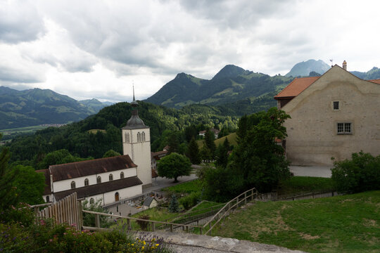 Gruyeres, Switzerland - 14 May 2026: View of the Eglise de Gruyeres church and historic buildings against a backdrop of lush green mountains under a cloudy sky.