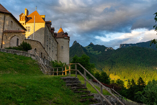 Gruyeres, Switzerland - 14 May 2026: View of Chateau de Gruyeres with its medieval stone walls and orange-tiled roofs overlooking lush green mountains under a dramatic sunset sky.