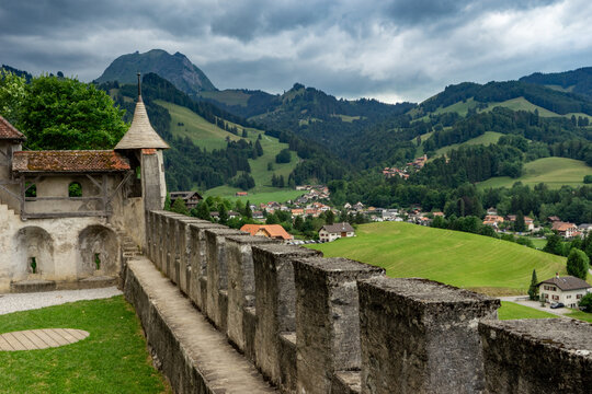 Gruyeres, Switzerland - 14 May 2026: View of Gruyeres Castle stone walls and crenellations overlooking the green valley, village houses, and the Swiss Prealps under a cloudy sky.