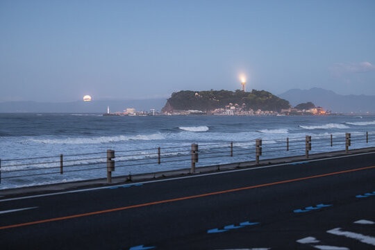 View of Shichirigahama Beach with waves crashing near the road and Enoshima island with its lit lighthouse under a full moon at dusk Kamakura, Kanagawa, Japan.